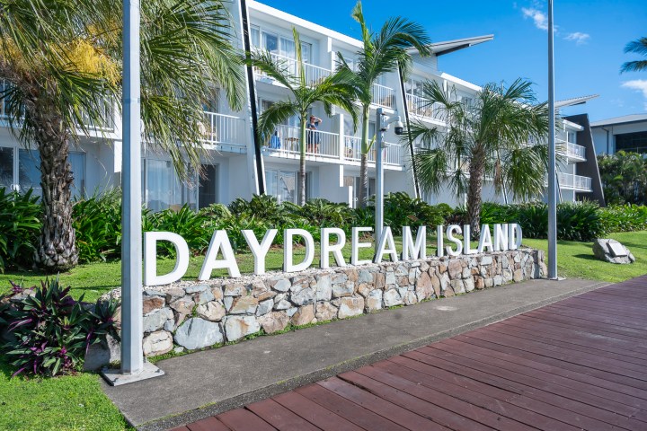 Sign reading 'Daydream Island' with white buildings and palm trees in the background.