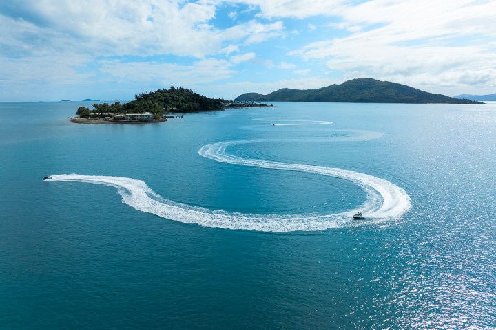 Aerial view of a jet ski creating curving white trails in blue ocean near a small island.