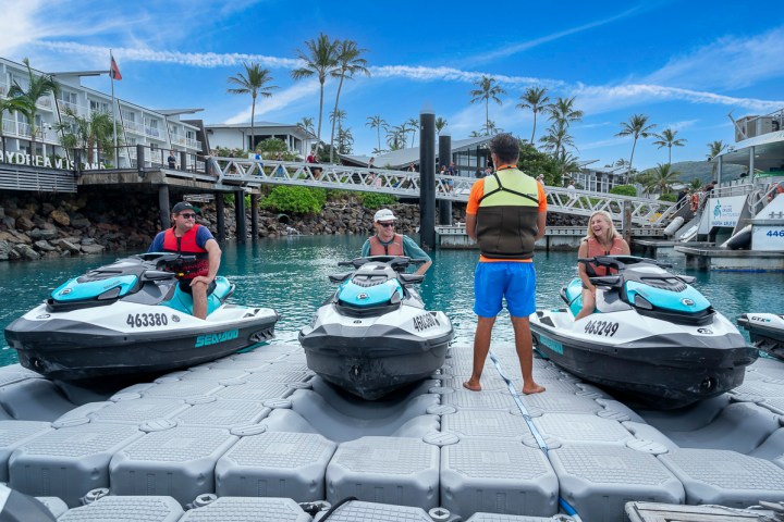 Three people on jet skis receiving instructions from a guide on a floating dock by palm trees.