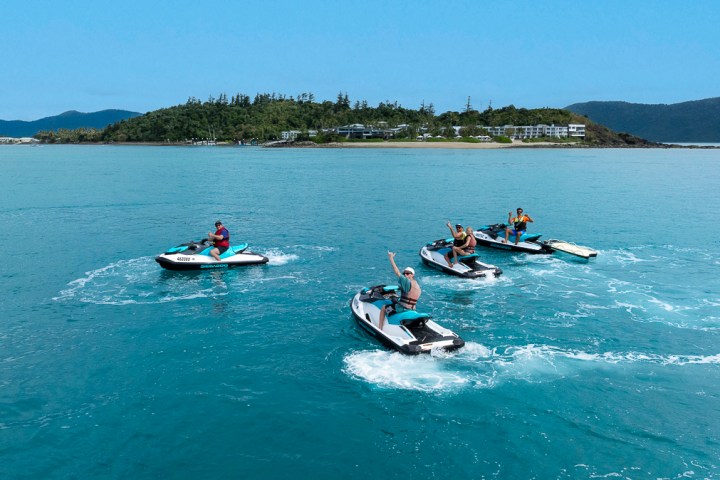 Four people on jet skis in turquoise water near an island with buildings and greenery.