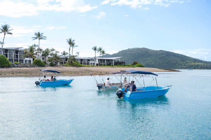 Three small blue boats with people, near a beach with palm trees and buildings in the background.