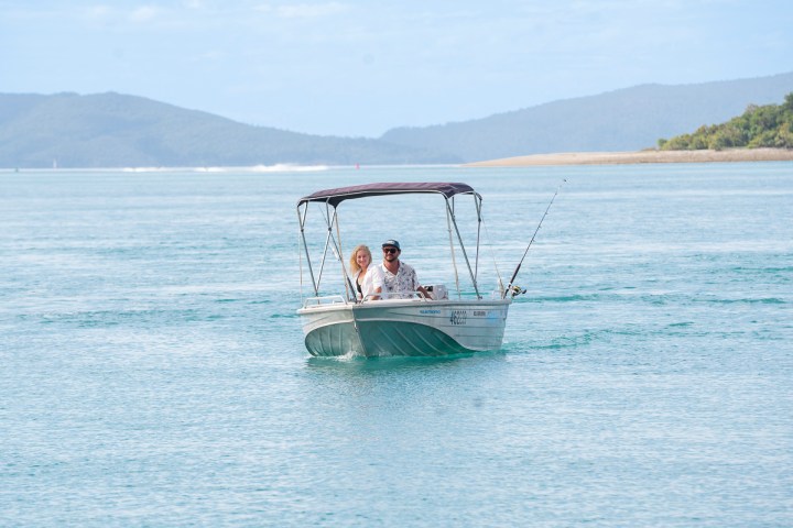 Couple on a small boat with a canopy, fishing on calm water, hills in background.