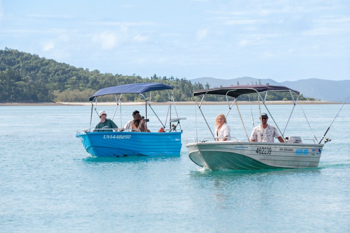 Two small motorboats with canopies on calm water, each carrying two people, with forested hills in the background.