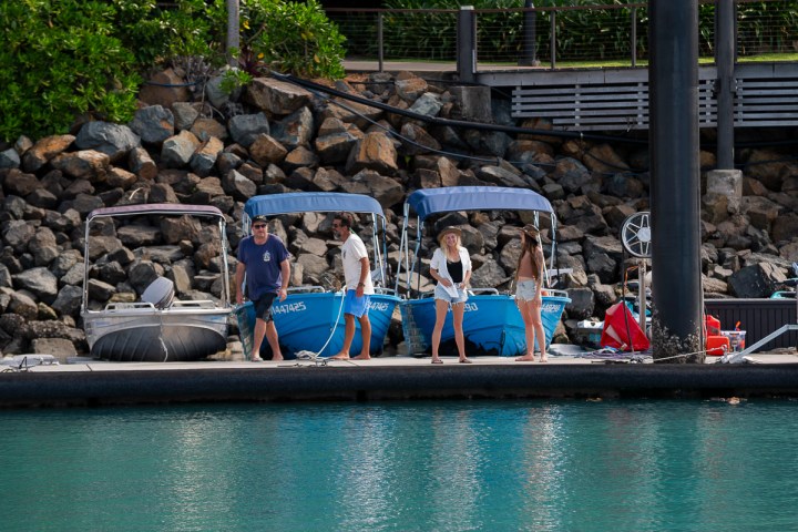 Four people standing on a dock with two small boats and rocky background.