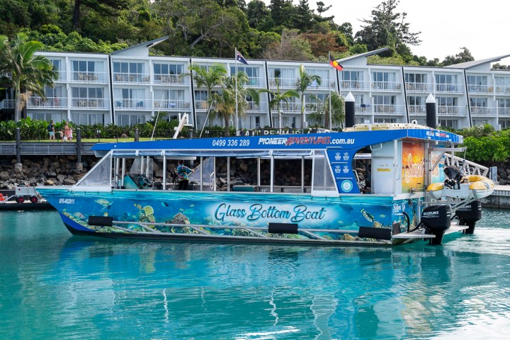 Glass bottom boat on water near hotel with palm trees and balconies.