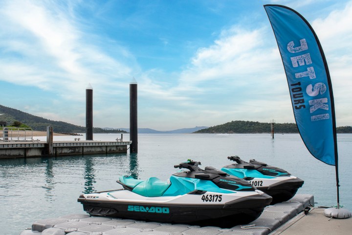 Two jet skis on a dock with a 'Jet Ski Tours' flag by a calm sea.