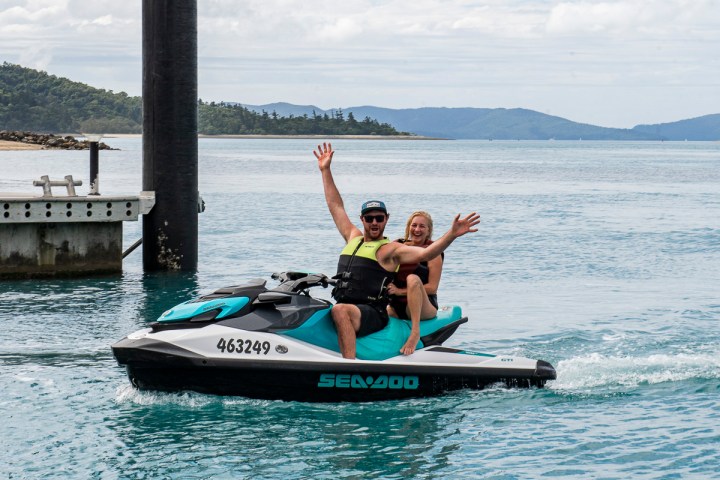 Two people joyfully riding a jet ski near a dock with a scenic backdrop of water and hills.