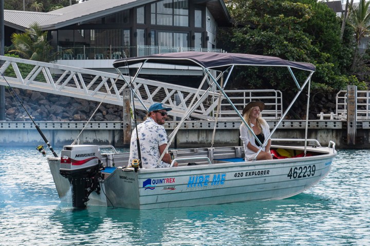 Two people on a small motorboat labeled 'Hire Me' in a marina setting.