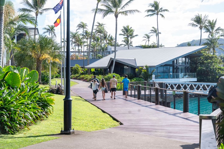Four people walking on a boardwalk by tropical trees and modern building.