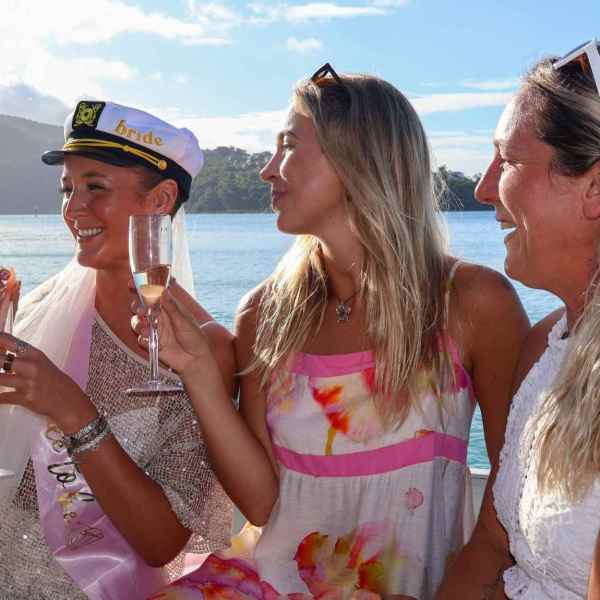 Three women on a boat, one wearing a captain's hat labeled 'bride', holding drinks, with ocean and island backdrop.