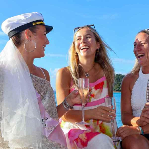 Three women laughing on a boat, one wearing a captain's hat and veil, holding champagne glasses.