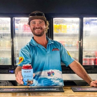 Man in cap smiling, holding a drink behind a bar fridge.
